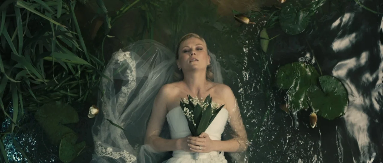 Melancholia (2011) movie still: medium — A woman in a white wedding gown lies flat on her back in a dark pond. She holds a bouquet of white flowers…
