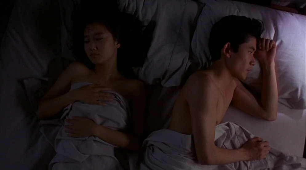 Mystery Train (1989) movie still: medium — A man and a woman lie together in a bed with soft grey sheets. They are both awake but turned away from…