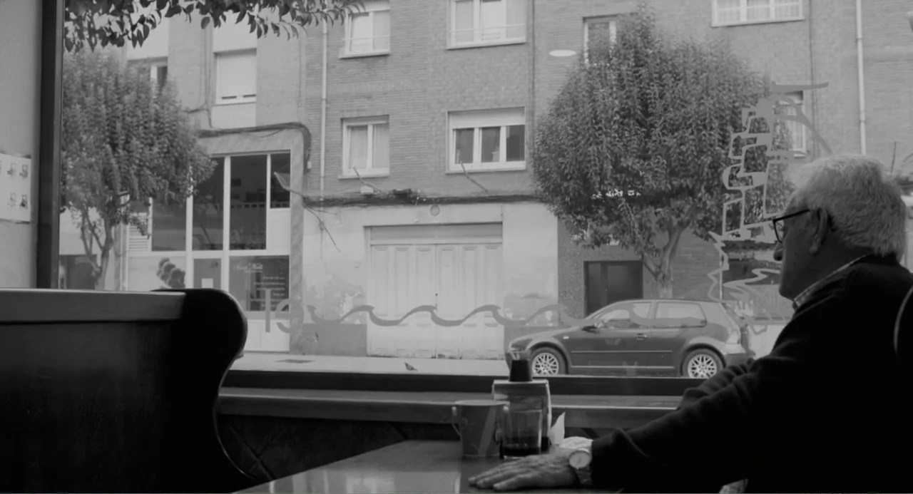 El Planeta (2021) movie still: medium — An elderly man sits at a cafe table looking out through a large window. Beyond the glass, a quiet street…