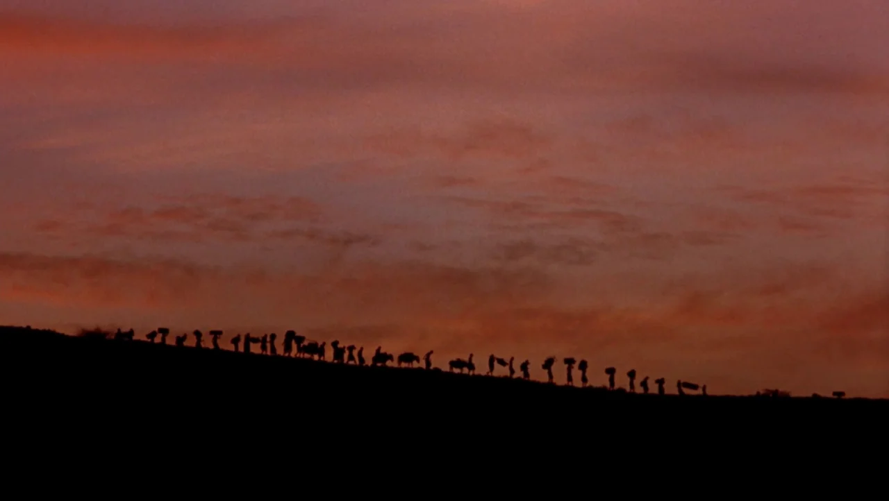 Mountains of the Moon (1990) movie still: wide — A long line of people in silhouette traverses a ridge against a deep red sky. The group carries…