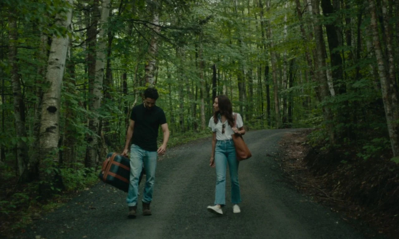 Black Bear (2020) movie still: fullbody — A gravel road winds through a lush, dense forest with towering deciduous trees. A man carrying a suitcase…