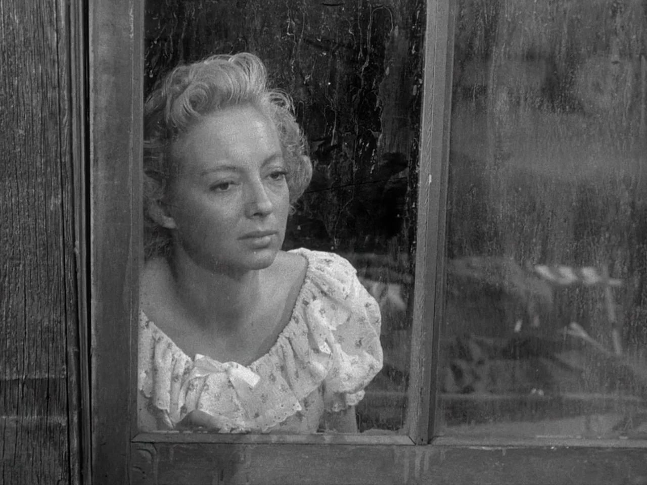 The Prowler (1951) movie still: closeup — A woman looks through a smudged and weathered glass window pane. She wears a floral blouse while gazing…
