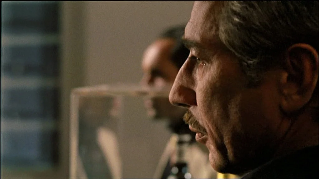 Chinese Roulette (1976) movie still: closeup — An interior space with a blurred figure in the background behind a glass partition. The man in the…