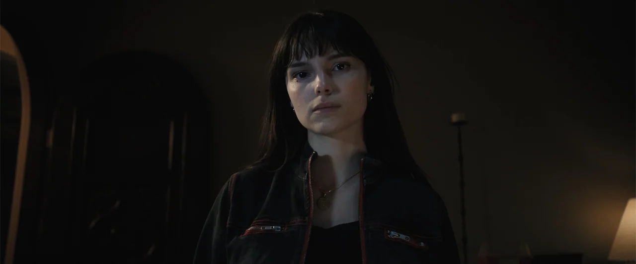 Trap House (2025) movie still: medium — A dimly lit interior space with a dark doorway visible in the background. A young woman stands looking…