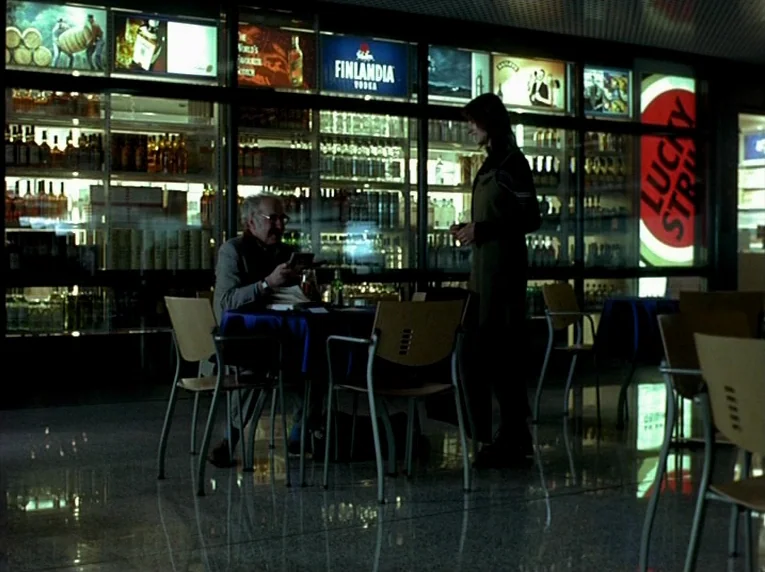 Notre Musique (2004) movie still: medium — A quiet airport-style shop is lined with rows of backlit liquor and cigarette displays. An older man sits…