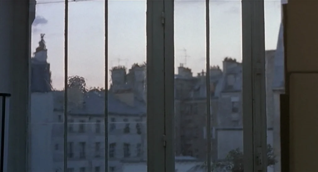 Vendredi Soir (2002) movie still: establishing — A view through a white-framed window looking out onto a densely built urban landscape. Multi-story…