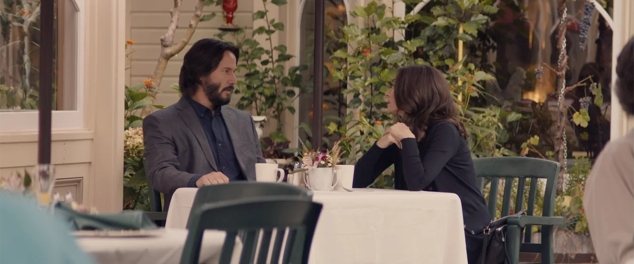 Destination Wedding (2018) movie still: medium — A man and a woman sit at a white-clothed table in a garden setting. They are engaged in…