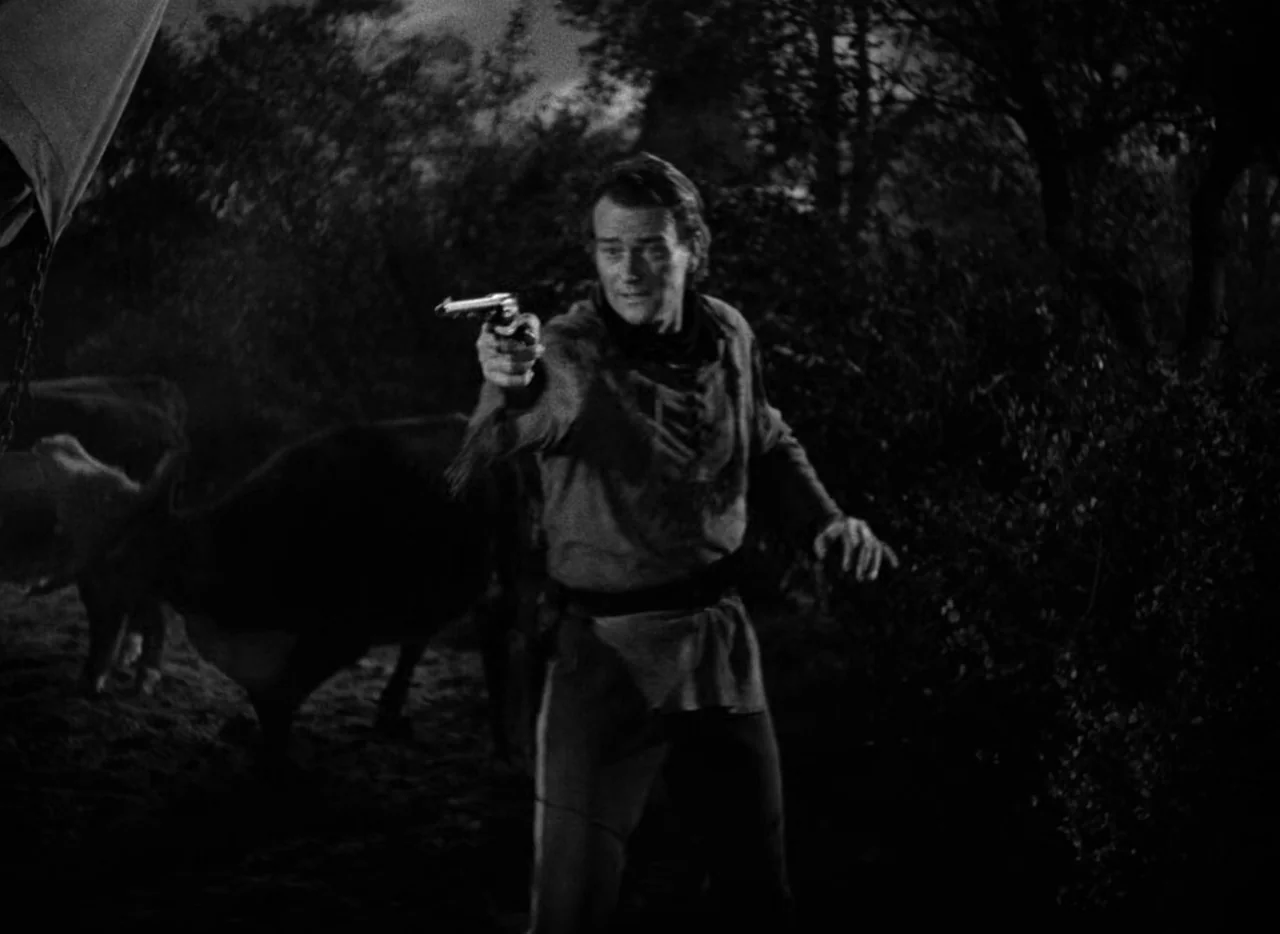 Red River (1948) movie still: fullbody — A nighttime encampment features dark, indistinct trees and livestock grazing in the shadows. A man stands…