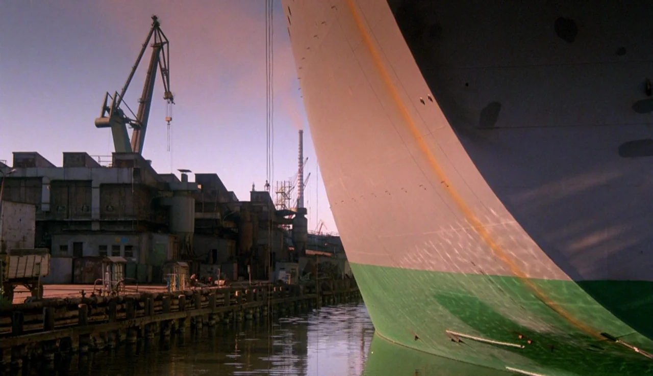 The Rainbow Thief (1990) movie still: establishing — A large ship hull occupies the right side of the frame at a shipyard. Industrial cranes and…