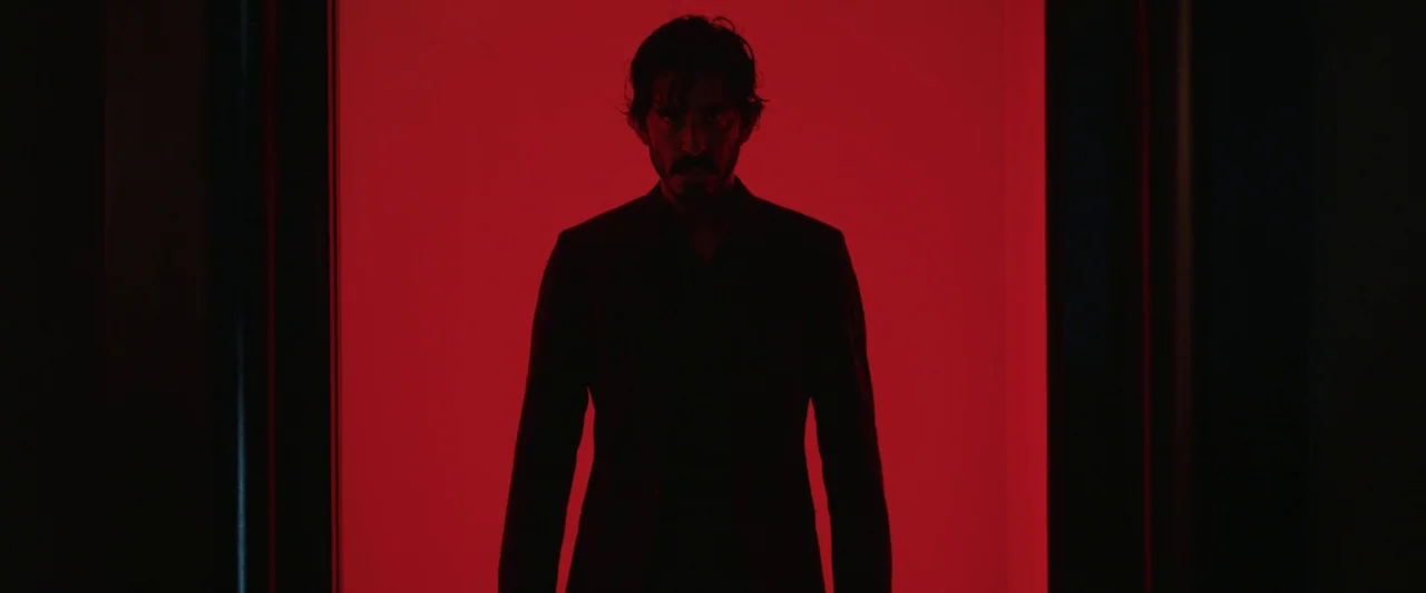 Monkey Man (2024) movie still: medium — A man stands in a dark doorway framed by a solid, bright red background. The subject is silhouetted against…