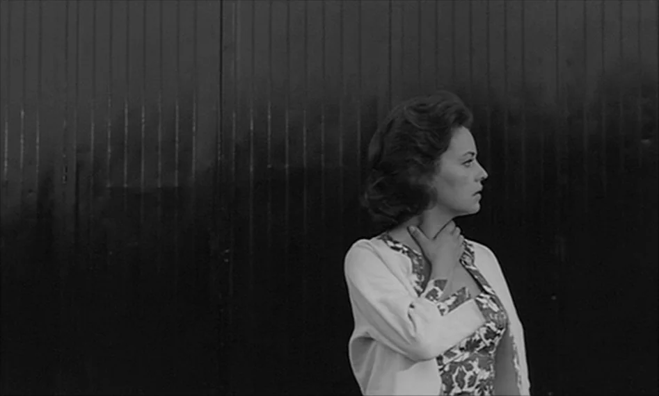 La Notte (1961) movie still: medium — A woman stands in front of a tall, dark vertical-slatted metal or wooden gate. She is looking off-screen to…