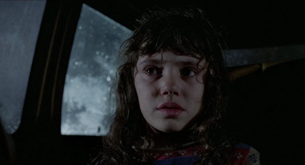 The Amityville Horror (1979) movie still: closeup — A young child sits inside a dark vehicle on a rainy night. She stares out the window while…