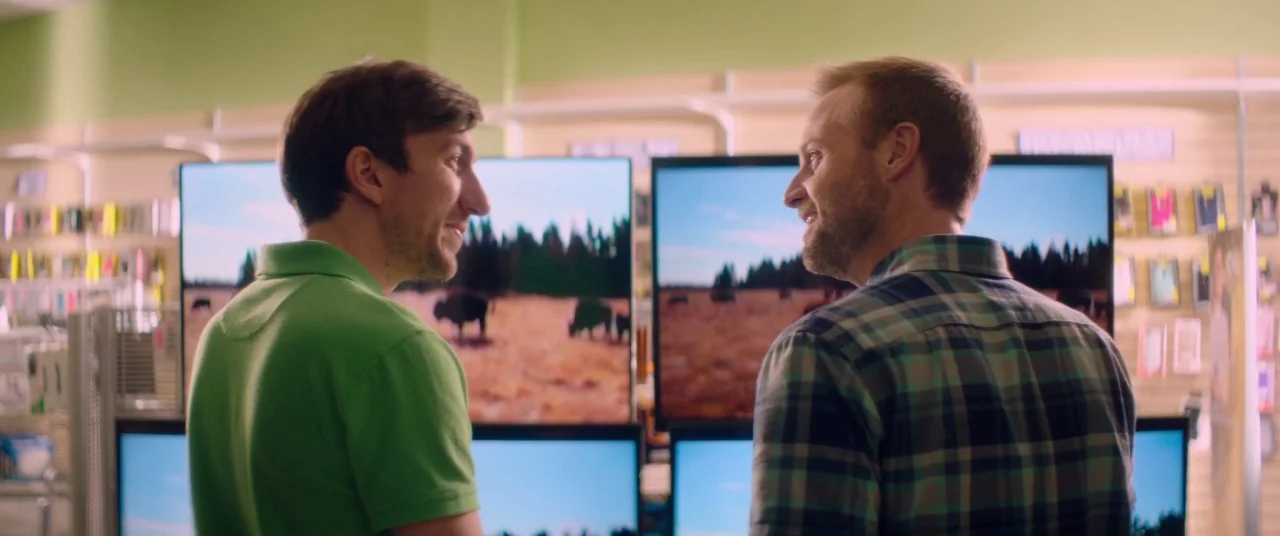 Room For Rent (2017) movie still: medium — A retail floor features walls lined with numerous large televisions displaying landscapes. Two men face…