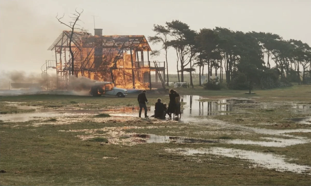 The Sacrifice (2015) movie still: wide — A flooded coastal meadow featuring a burning two-story house frame and a smoke-filled sky. A small group of…