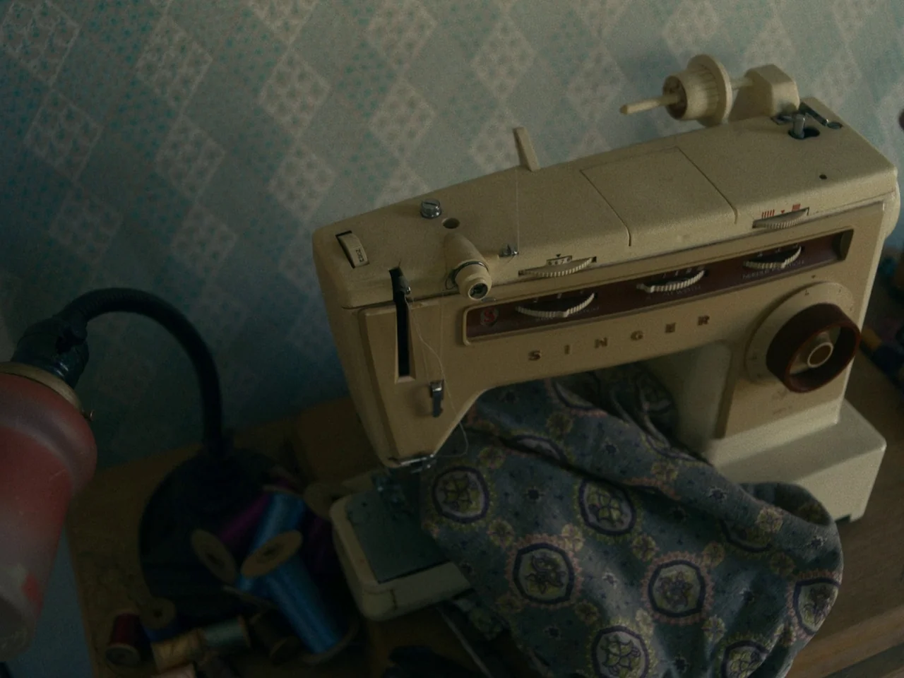 I’m Thinking of Ending Things (2020) movie still: closeup — A vintage beige Singer sewing machine sits on a wooden desk against blue patterned…