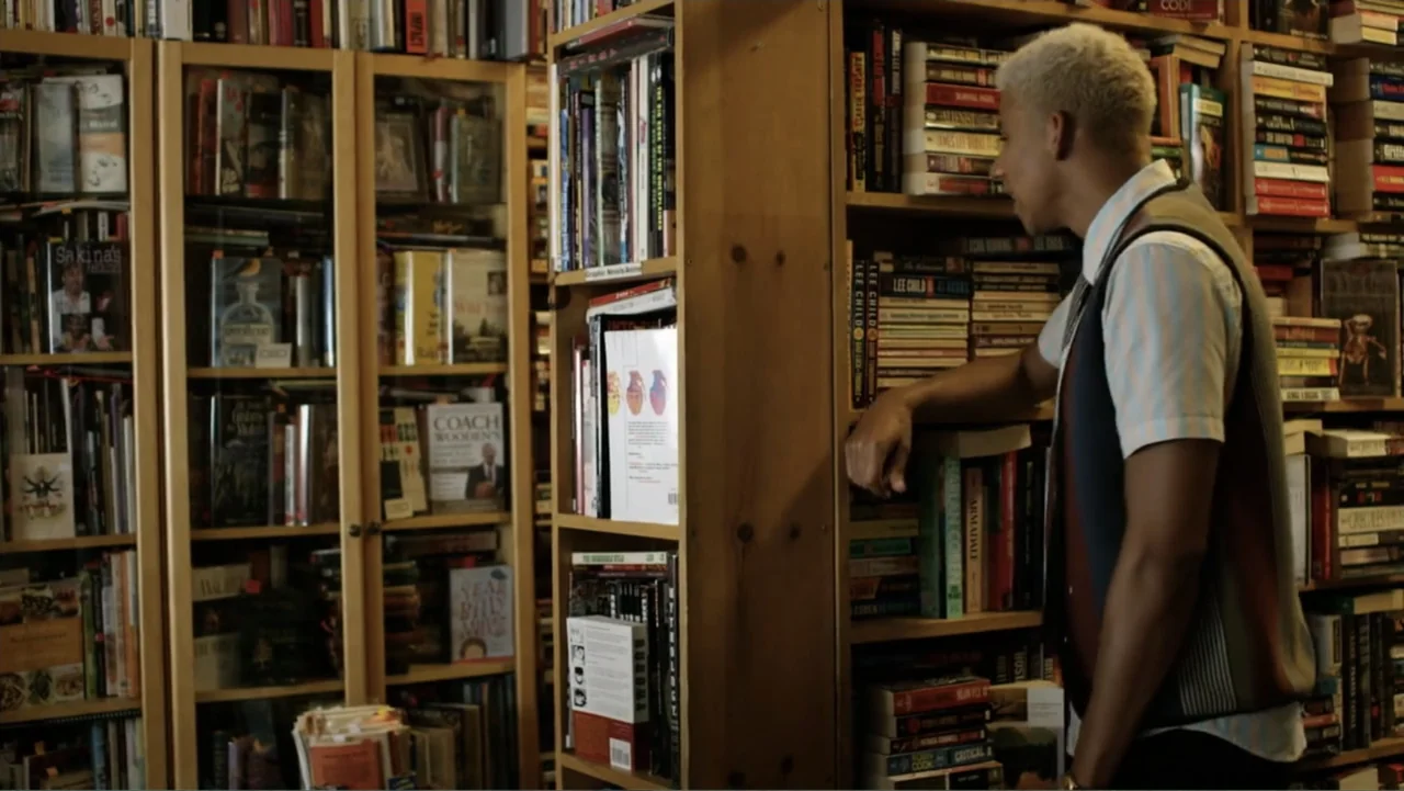 Like.Share.Follow. (2017) movie still: medium — A dense bookstore aisle features wooden shelves tightly packed with books of varying sizes. A young…