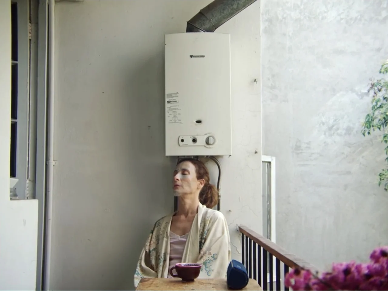 History and Geography (2024) movie still: medium — A woman sits on a narrow balcony against a plain white wall. She wears a floral robe and has a…