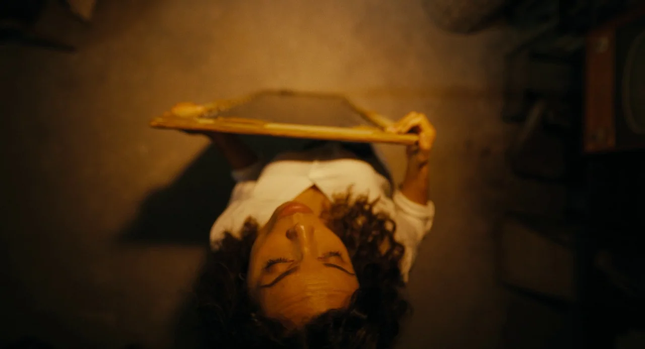 Barbarian (2022) movie still: medium — A woman stands in a dimly lit room with brown concrete flooring. She holds a framed rectangular mirror…