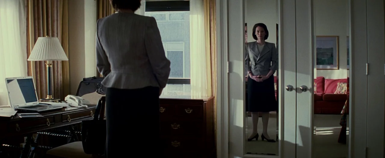 Michael Clayton (2007) movie still: medium — A hotel room interior contains a wooden desk with a laptop and a wardrobe mirror. A woman stands in…