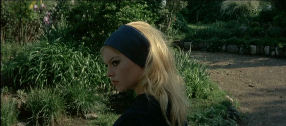 Contempt (Le Mépris) (1963) movie still: medium — A woman stands in a lush outdoor area with dense green foliage and shrubs. She turns her head in…