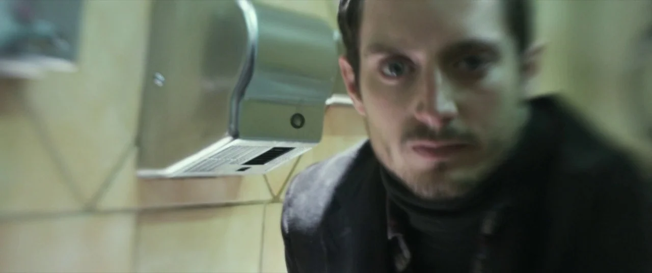 Maniac (2012) movie still: closeup — A public washroom featuring tan tile walls and a silver metal hand dryer. A man faces the camera directly while…