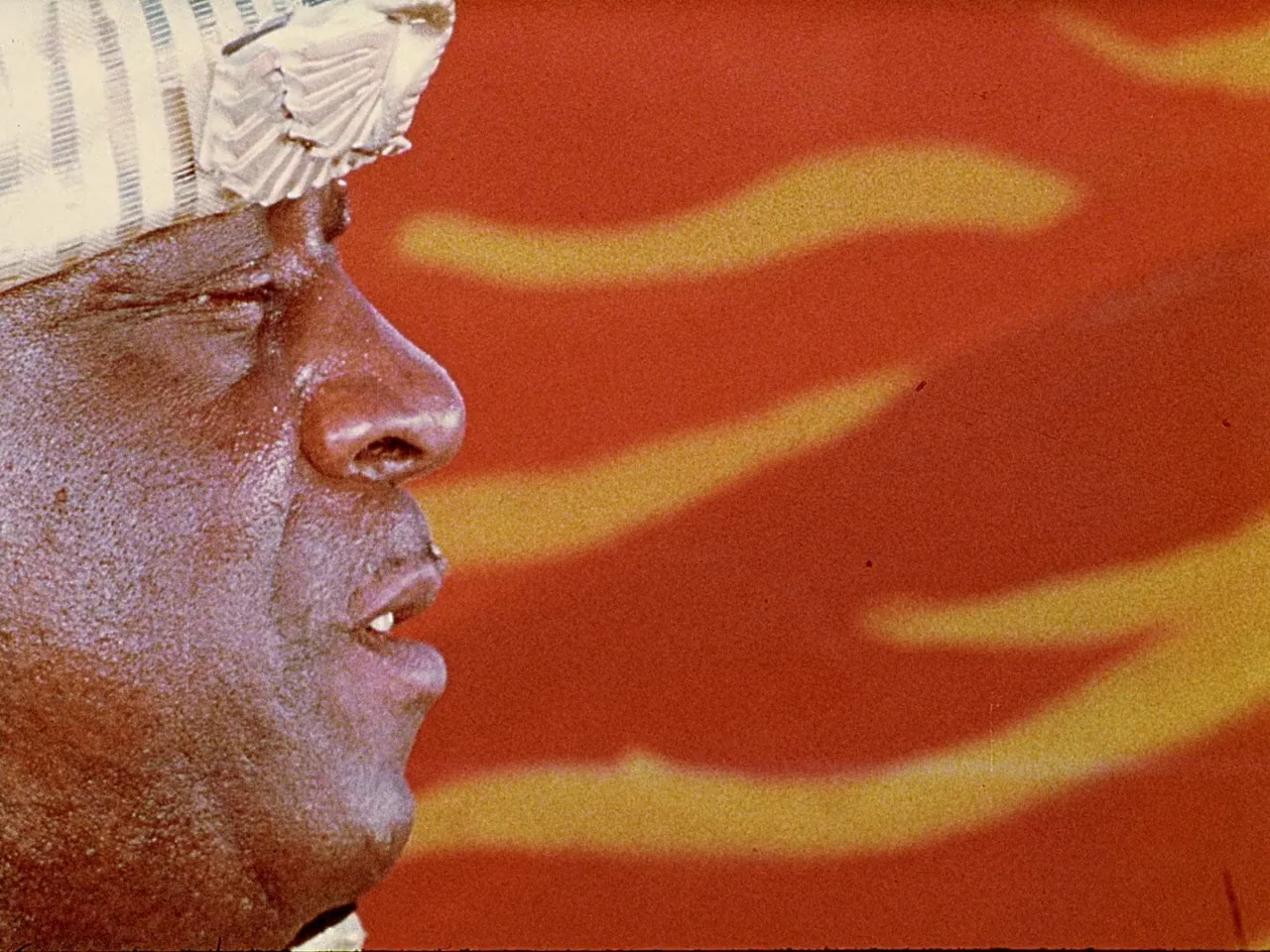 Space is the Place (1974) movie still: closeup — A close-up shot of an older Black man wearing a striped hat. He is outdoors in front of a red and…
