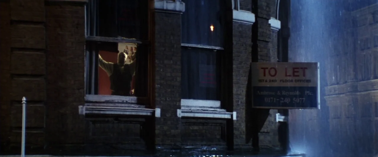 Mission: Impossible (1996) movie still: wide — A dark brick building at night features a glowing window and a rental sign. A man stands in the open…