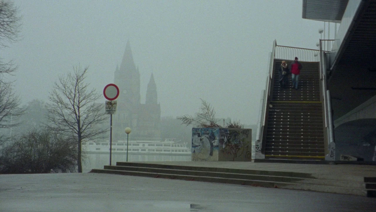 Museum Hours (2012) movie still: wide — An outdoor staircase leads up to a pedestrian bridge under a dense, foggy sky. A pair of people descend the…