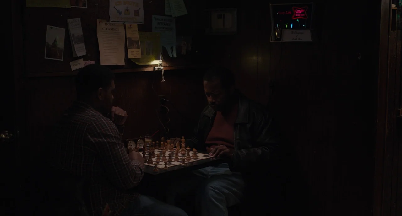 Paterson (2016) movie still: medium — Two men sit at a small table playing a game of chess in a dark tavern. The wood-paneled wall behind them is…