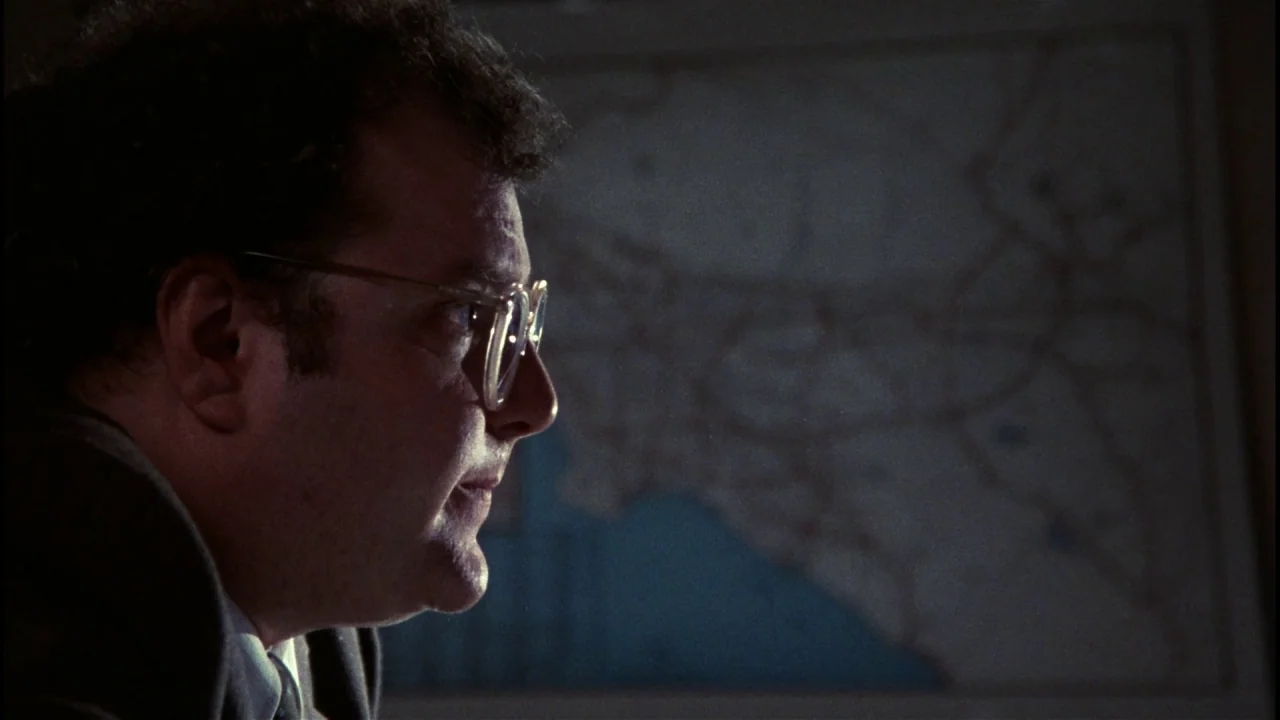 Star 80 (1983) movie still: closeup — A dark office space with a large wall map visible in the background. A man stands in the foreground, his…