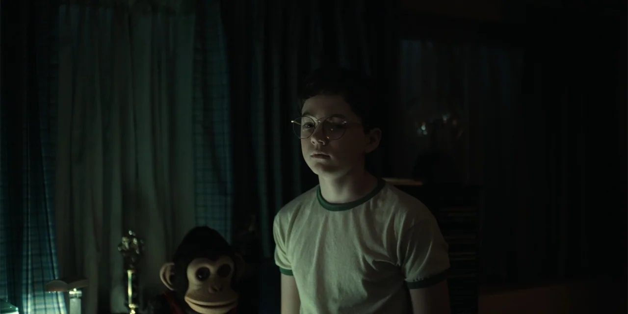 The Monkey (2025) movie still: medium — A dimly lit room with heavy curtains and a toy monkey on a shelf. A young boy in a t-shirt stands positioned…