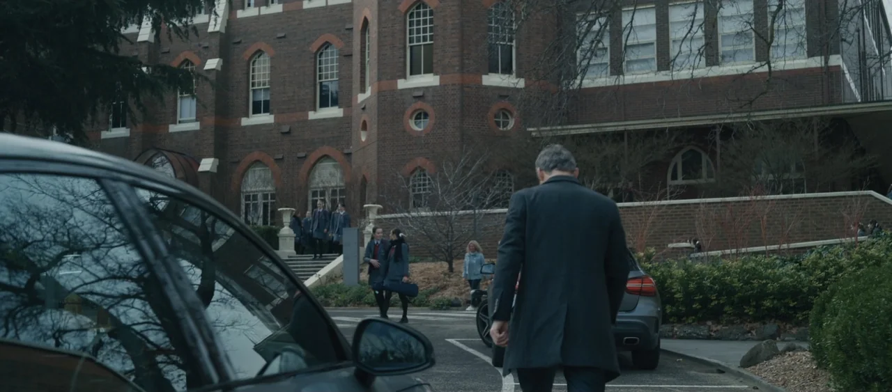 Force of Nature: The Dry 2 (2024) movie still: medium — A brick multi-story building stands behind a parking area with several parked cars. A…