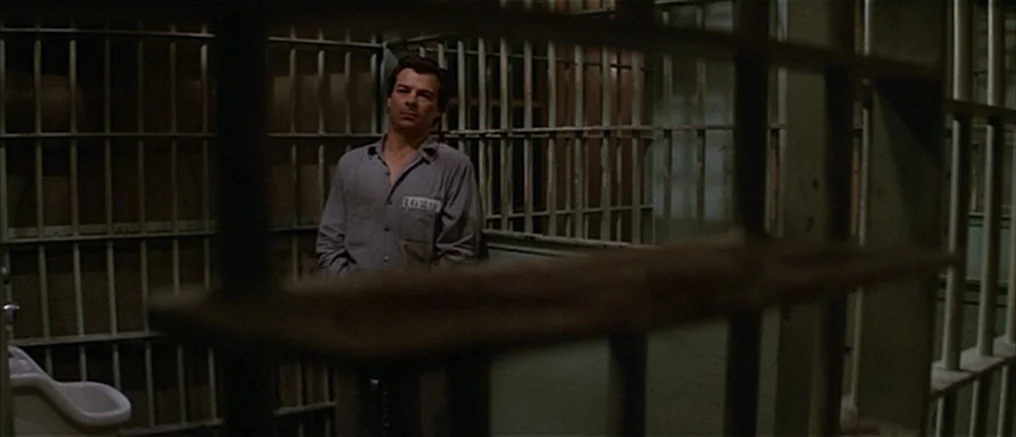 Assault on Precinct 13 (1976) movie still: medium — A dimly lit room defined by heavy vertical metal bars surrounding the area. A lone man in prison…