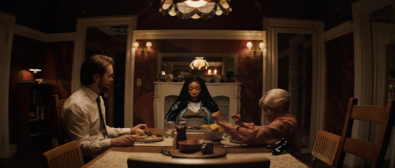 The Front Room (2024) movie still: wide — A dimly lit dining room features wood paneled walls and a mantelpiece fireplace. Three people sit at the…