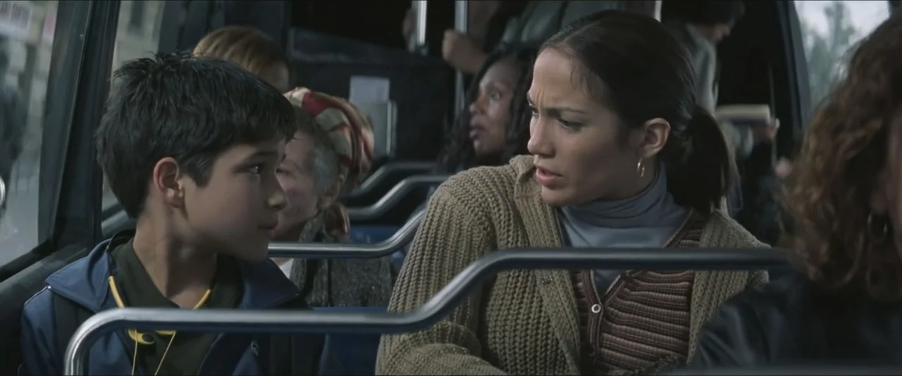 Maid in Manhattan (2002) movie still: medium — Passengers sit in the interior of a public transit vehicle with metal safety rails. A woman speaks to…