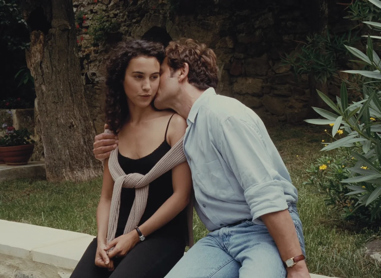 Autumn Tale (1998) movie still: medium — A stone courtyard with overgrown vegetation and old masonry walls frames two seated people. A man leans…