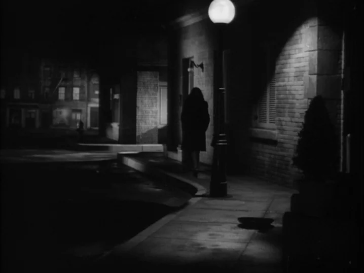 The Seventh Victim (1943) movie still: wide — A dark night on a concrete sidewalk beside a tall brick building. A lone figure walks away from the…