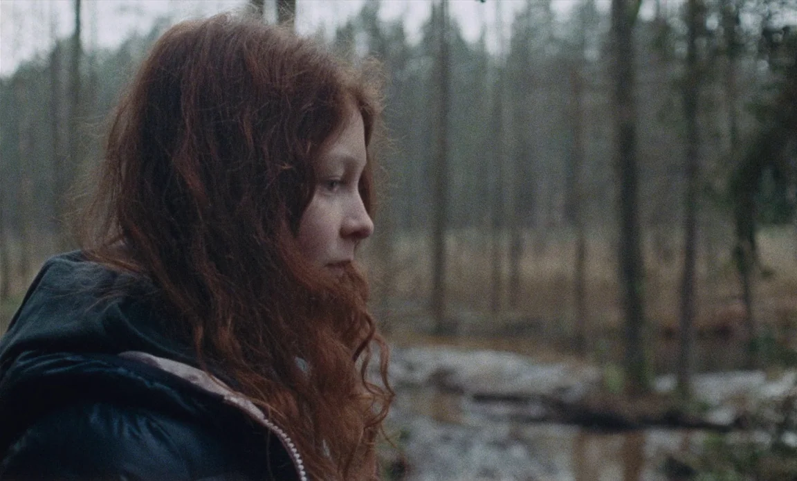 High Life (2018) movie still: medium — A young woman with auburn hair stands in a sparse woodland during daytime. The background shows thin leafless…
