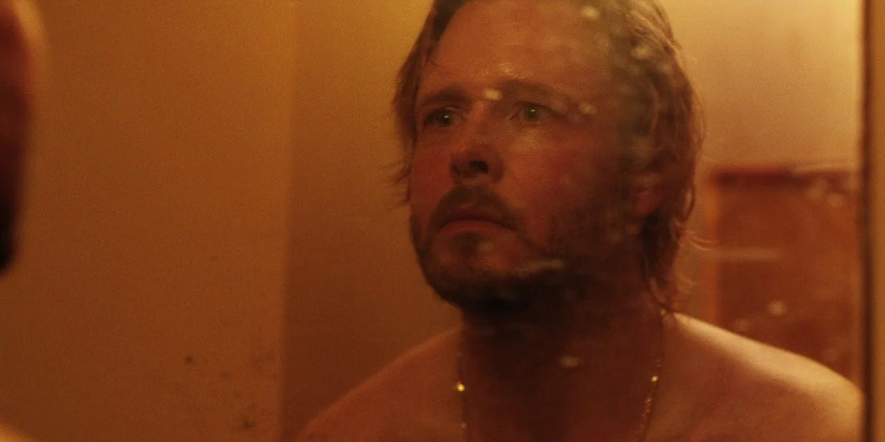 The Death of Dick Long (2019) movie still: closeup — A dimly lit room seen through a reflection in a spotted mirror. A man stares directly at his…
