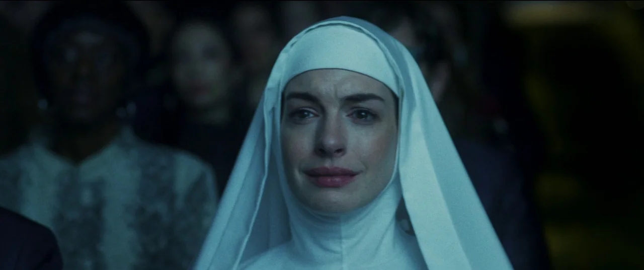 She Came To Me (2023) movie still: closeup — A dimly lit indoor space with blurred figures present in the background. A woman dressed in a nun's…