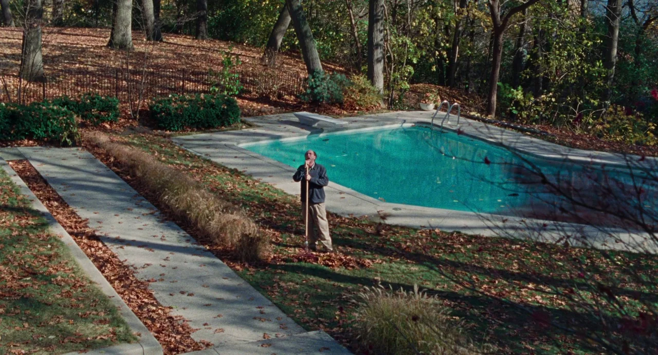 Dream Scenario (2023) movie still: wide — A man stands on a residential lawn littered with brown fallen autumn leaves. He grips a tall rake while…