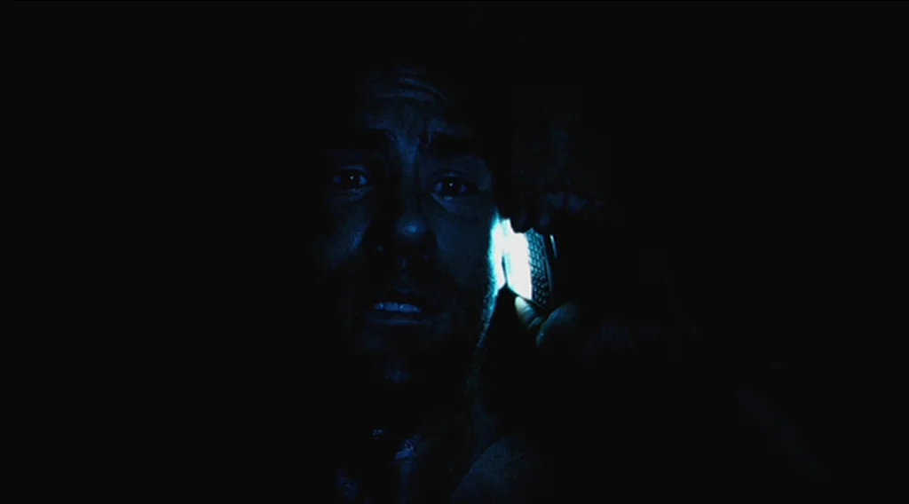 Buried (2010) movie still: closeup — A pitch-black, enclosed space with limited visibility. A man's face is illuminated by a mobile phone held to…