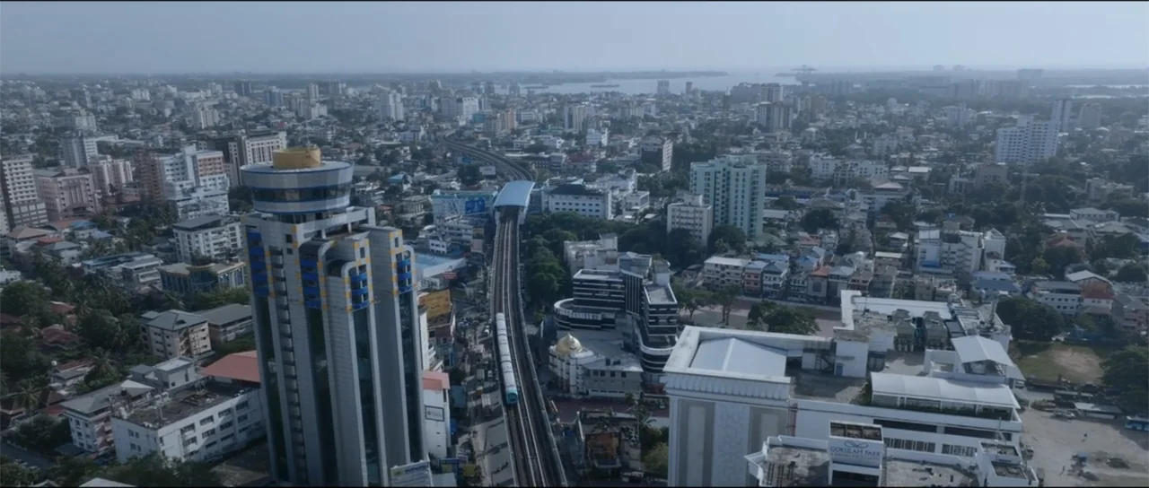 Garudan (2024) movie still: establishing — A wide aerial view shows a dense city with high-rise buildings and transit lines. A metro train travels…