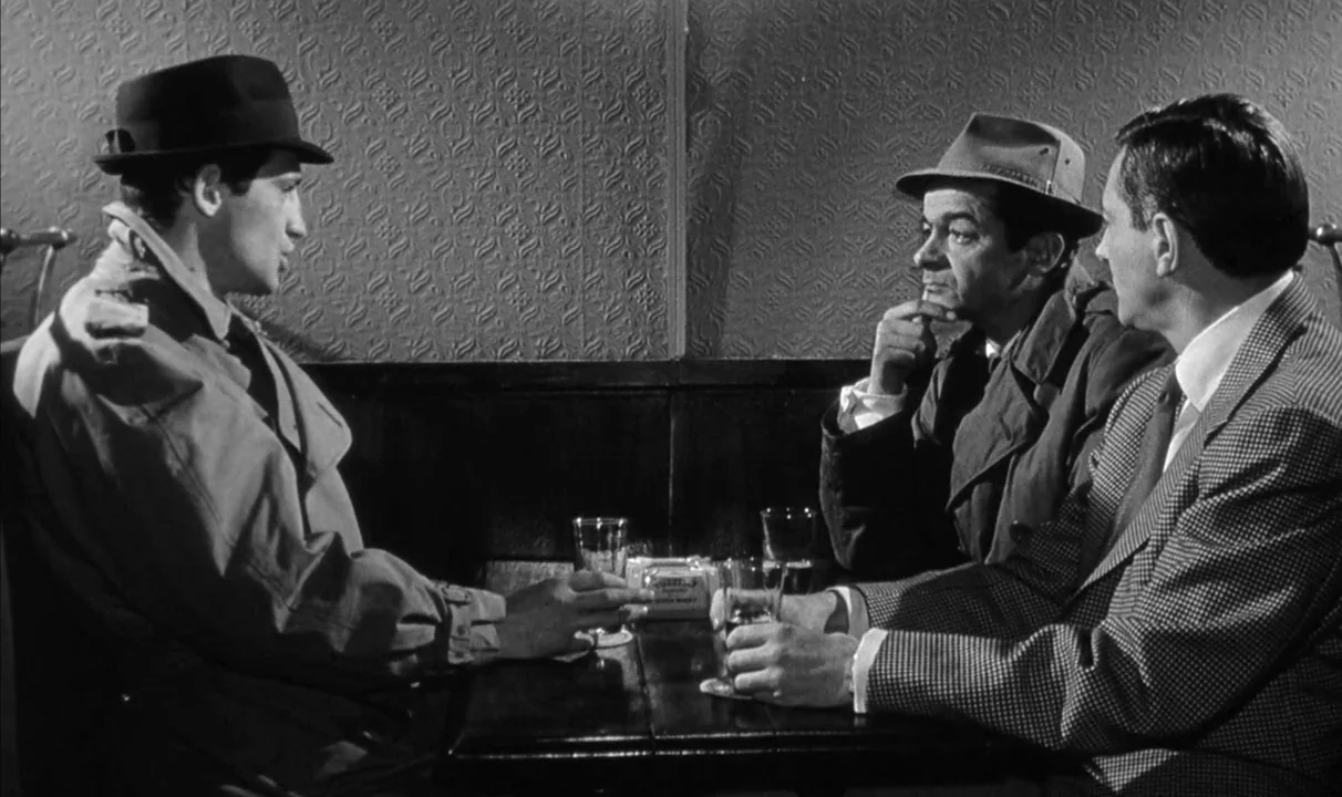 Le Doulos (1962) movie still: medium — Three men are seated together at a small table inside a dimly lit booth. They sit in conversation against a…