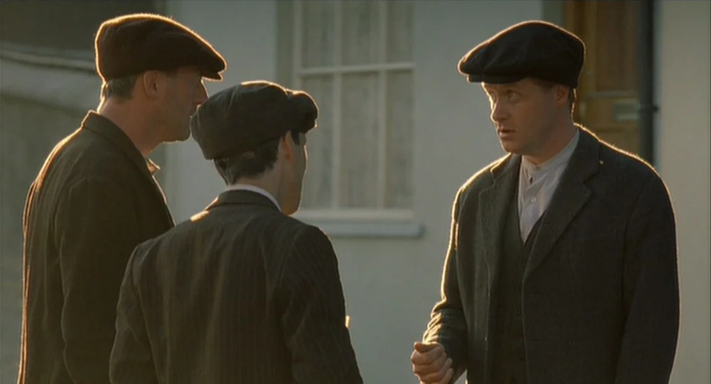 The Wind That Shakes the Barley (2006) movie still: medium — Three men in early 20th-century attire gather outside a plain, light-colored building.…