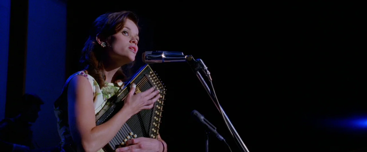 Walk The Line (2005) movie still: medium — A dark concert venue with a performer illuminated by a tight blue spotlight. A young woman in a floral…