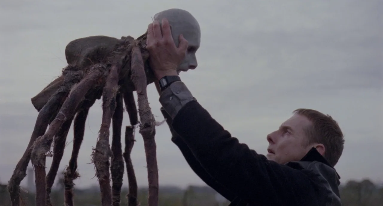 Possum (2018) movie still: medium — An overcast sky hangs above a vast, open field. A man raises a large, spindly, multi-legged construct with a…