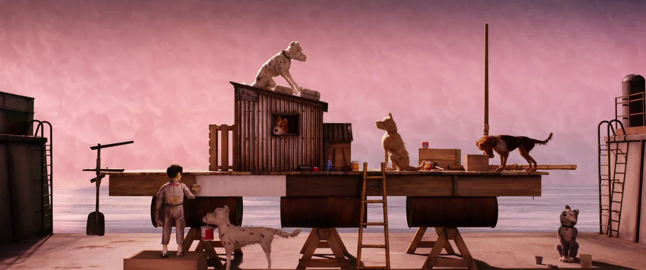 Isle of Dogs (2018) movie still: wide — A wooden platform raised on barrels sits on a dock against a pink sky. A young boy paints a white stripe on…
