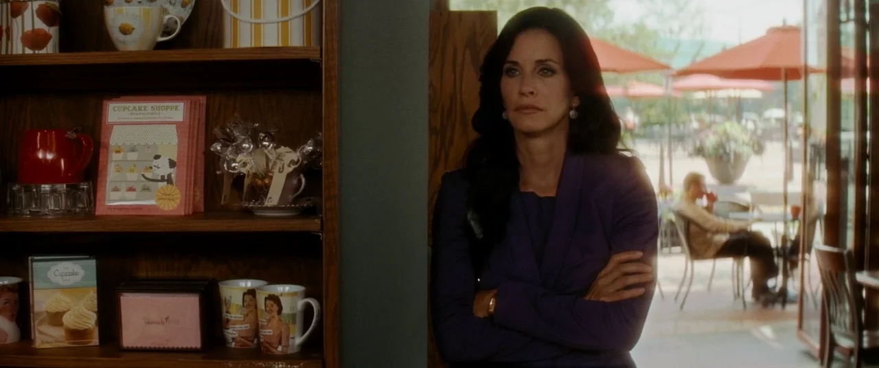 Scream 4 (2011) movie still: medium — A retail gift shop features wooden shelves displaying mugs and small stationery items. A woman in a purple…