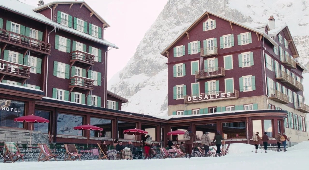 Phantom Thread (2017) movie still: wide — Two large connected burgundy-colored hotels with green shutters sit against a snowy mountain backdrop.…